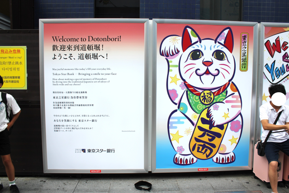 道頓堀の街頭に設置された巨大パネルの浮世絵風招き猫のクローズアップ画像。白の体に星や花柄、浮雲などの意匠が描かれている。ポップな色と親しみやすい猫の表情が特徴