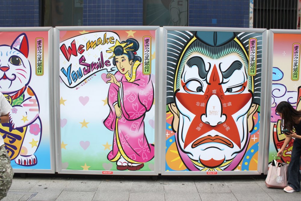 道頓堀の街頭に設置された巨大パネルの浮世絵風の女性と歌舞伎役者の顔のクローズアップ画像。ピンクの着物を着た女性が身をかがめるように微笑み、「We make You smile」という英語の文字と共に描かれた浮世絵風イラスト。背景はカラフルな星とハートでポップな雰囲気。歌舞伎風の男性の顔が大きく描かれた浮世絵風イラスト。顔の中央には赤い星型の模様があり、東京スター銀行を想起させる星柄が配してある。