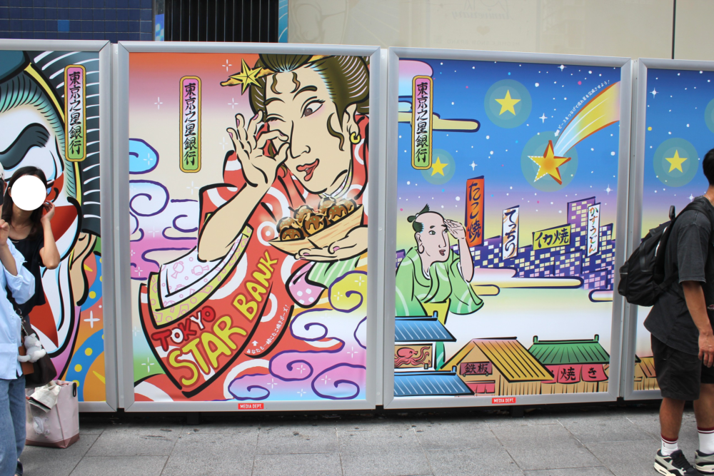 道頓堀の街頭に設置された巨大パネルの浮世絵風のたこ焼きを持った女性のクローズアップ画像。髪に星飾りをつけた着物姿の女性が、たこ焼きを手にして微笑む浮世絵風イラスト。背景は雲と虹色のようなグラデーションで、着物には「TOKYO STAR BANK」の文字が描かれている。