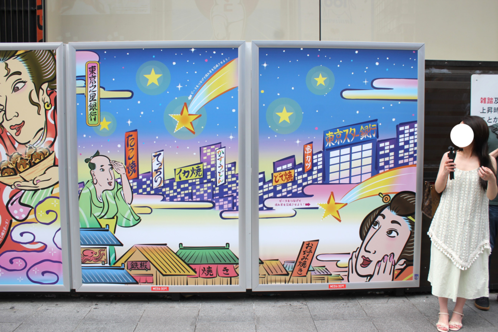 道頓堀の街頭に設置された巨大パネルの浮世絵風の大阪の夜景を描いたクローズアップ画像。夜空に大きな星や流れ星が輝く中、道頓堀の街並みが浮世絵風に描かれたイラスト。たこ焼きや焼きそばなどの看板が立ち並び、手前には驚いた表情の男性、右下には頬に手を添える女性が配置されている。