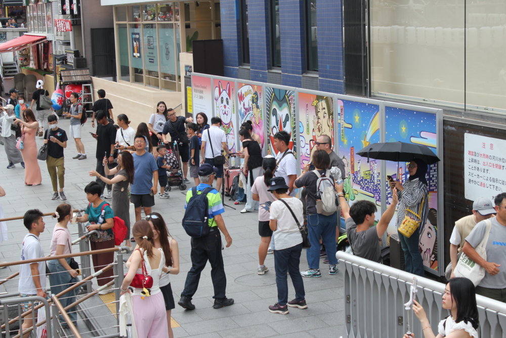 大阪・道頓堀の街中に設置されたカラフルな浮世絵風パネルの前を、多くの人々が行き交っている様子。アジア系の観光客が多く映っている