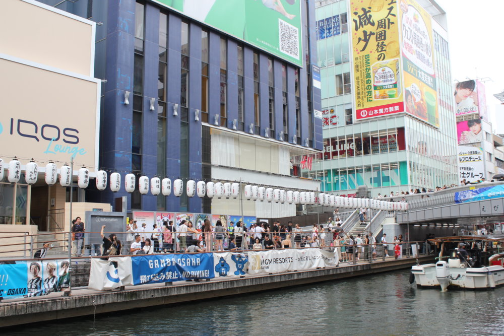 大阪・道頓堀の街中に設置されたカラフルな浮世絵風パネルの前を、多くの人々が行き交っている様子。。