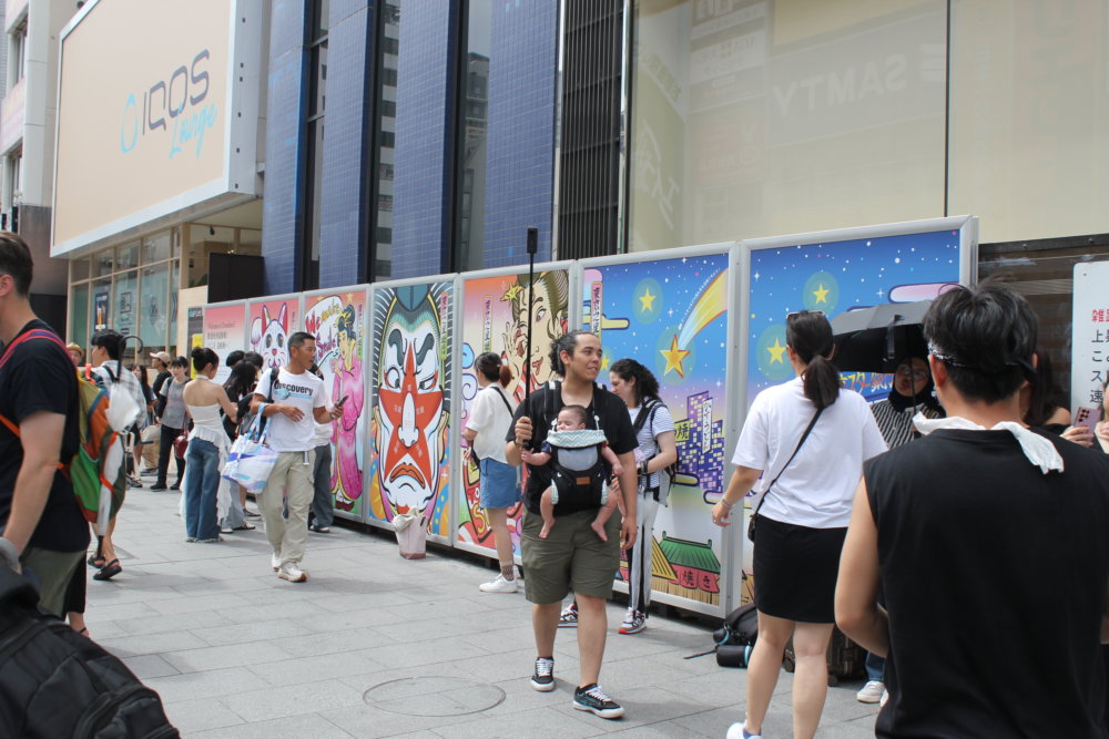 大阪・道頓堀の街中に設置されたカラフルな浮世絵風パネルの前を、多くの人々が行き交っている様子。パネルには招き猫、ピンクの着物の女性、歌舞伎風の顔、たこ焼きを持つ女性、星空と街並みのイラストが並んでいる。中央には赤ちゃんを抱っこ紐で抱えた人物が歩いており、周囲には観光客や買い物袋を持った人々が集まり、にぎわった雰囲気。