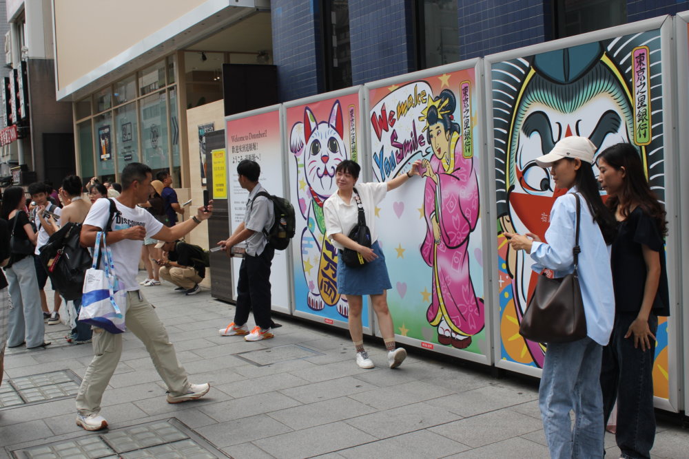 大阪・道頓堀の街中に設置されたカラフルな浮世絵風パネルの前を、多くの人々が行き交っている様子。パネルには招き猫、ピンクの着物の女性、歌舞伎風の顔、たこ焼きを持つ女性、星空と街並みのイラストが並んでいる。中央にはパネルと共に写真を撮る女性がいたりと、にぎわった雰囲気。
