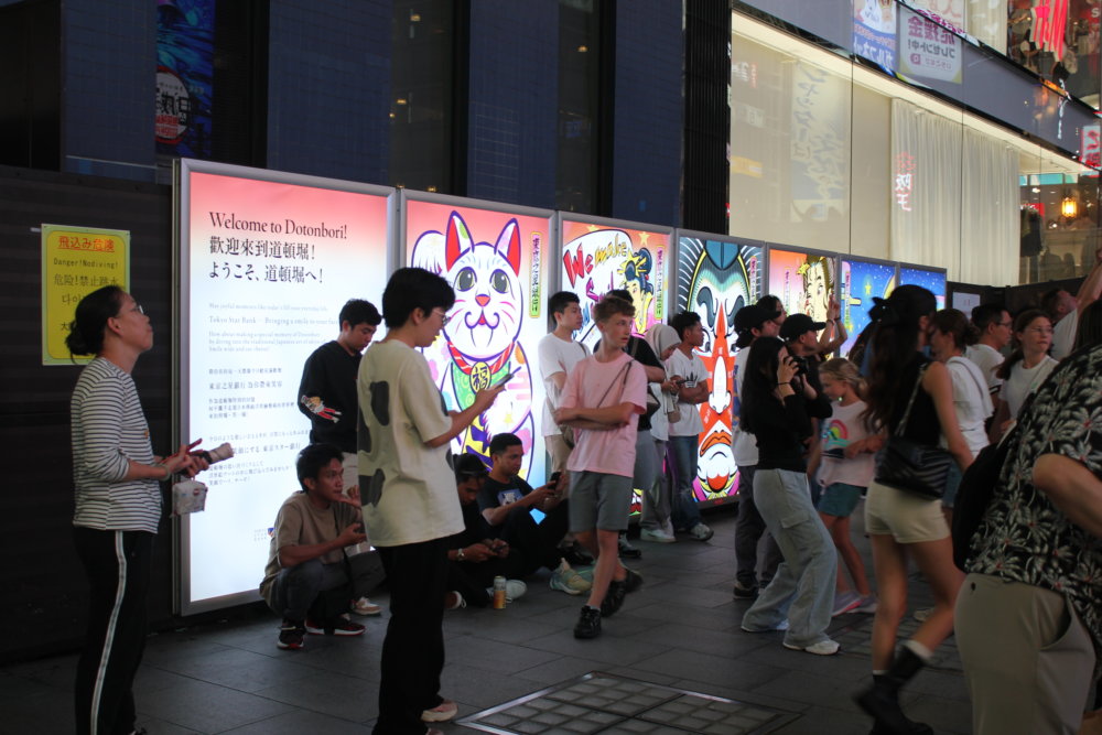 夜の風景 大阪・道頓堀の街中に設置されたカラフルな浮世絵風パネルの前を、多くの人々が行き交っている様子。アジア系の観光客が多く映っている。パネルは電光製で光り輝いている