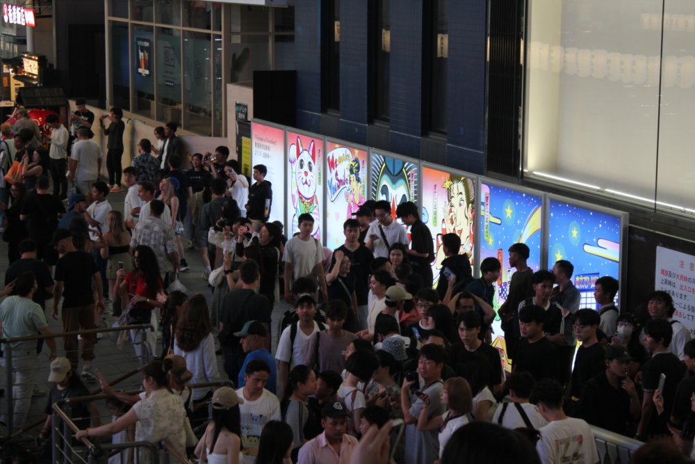 夜の風景 大阪・道頓堀の街中に設置されたカラフルな浮世絵風パネルの前を、膨大な数の人々が行き交っている様子。アジア系の観光客が多く映っている。パネルは電光製で光り輝いている