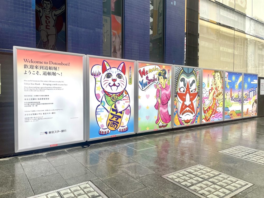 道頓堀の街頭に設置された巨大パネルに、浮世絵風のイラストが6枚並んでいる。左から招き猫、ピンクの着物姿の女性、歌舞伎風の顔の男性、たこ焼きを持った女性、大阪の江戸の街並みと現在の街並みが描かれており、カラフルでポップな色使い。