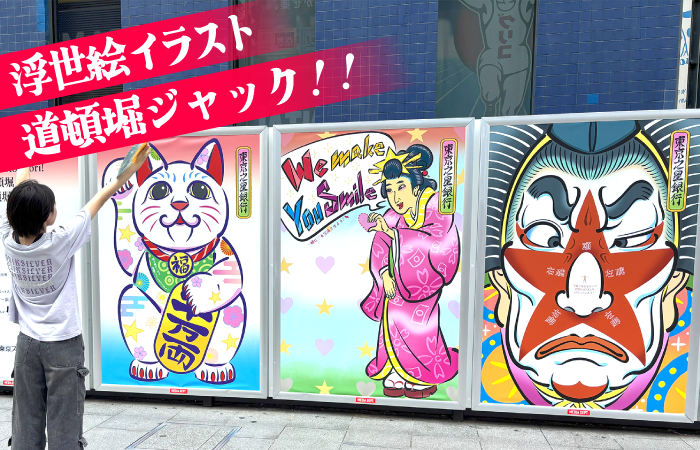 道頓堀の街頭に設置された巨大パネルに、浮世絵風のイラストが3枚並んでいる。左から招き猫、ピンクの着物姿の女性、歌舞伎風の顔の男性が描かれており、カラフルでポップな色使い。手前には白いTシャツの人物が手を上げて、イラストに触れるようなポーズを取っている。画面上部にはピンクの帯に「浮世絵イラスト 道頓堀ジャック!!」の文字。